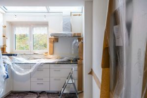 A kitchen in the middle of a renovation with protective plastic covering the cabinets and a ladder in the room.